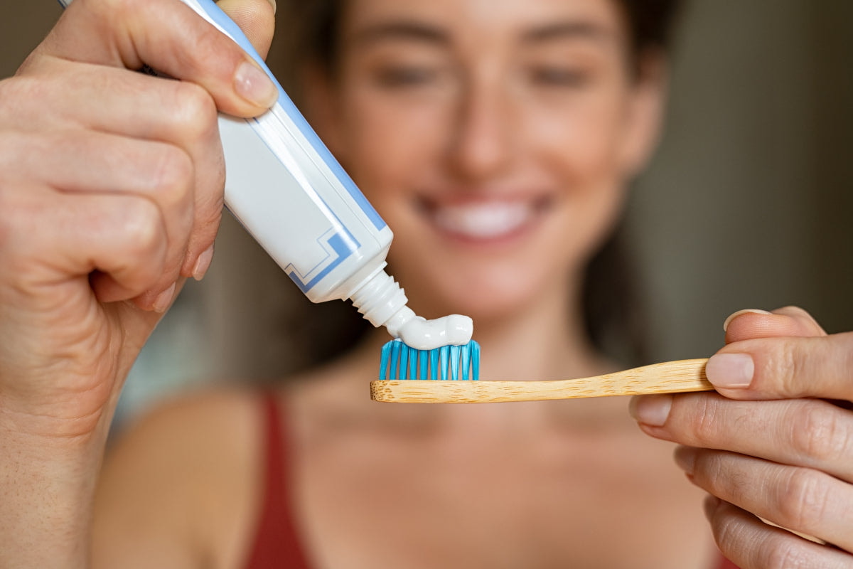 Close-up of a smiling woman squeezing toothpaste onto a bamboo toothbrush with blue bristles, holding both items in her hands.