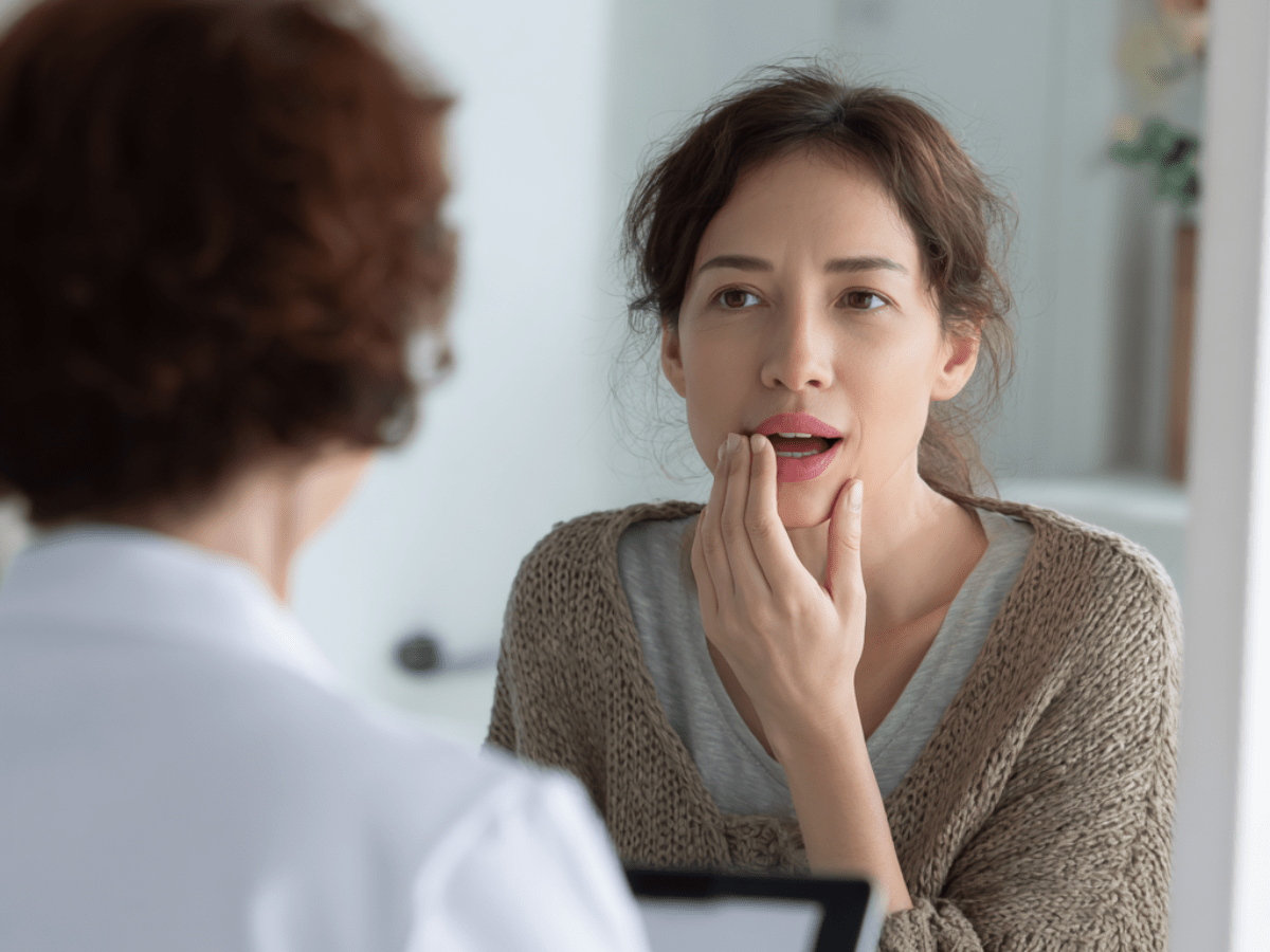 Young woman touching her jaw and describing oral discomfort to a female dentist during a consultation, possibly indicating toothache, gum pain, or jaw sensitivity