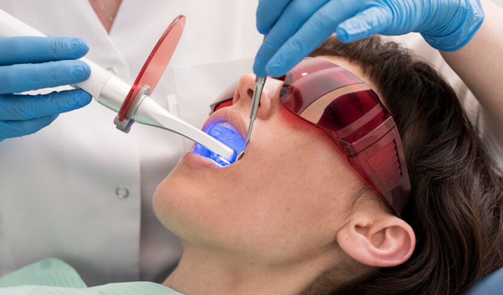 A woman sits in a dental chair while a dentist cleans her teeth with dental tools. 