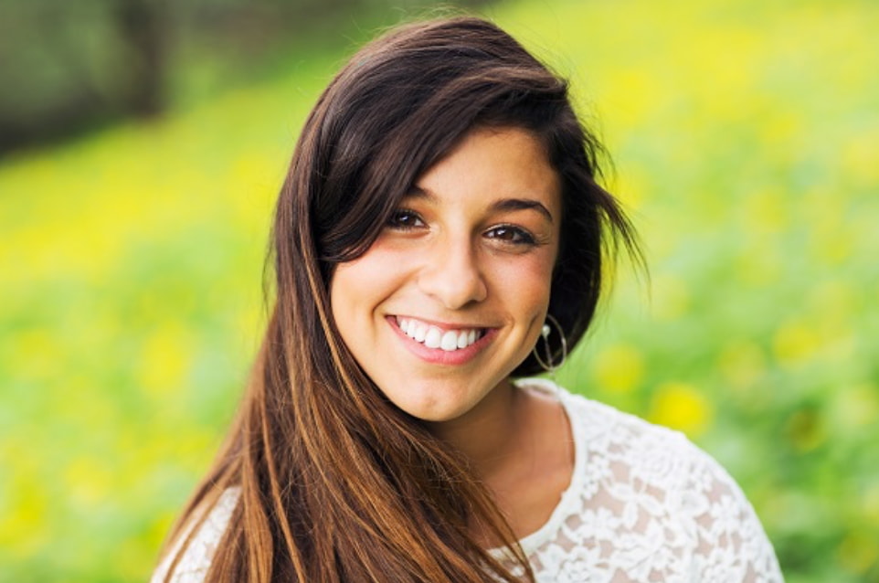 A young woman with long, dark hair stands in a flower-filled field, wearing a white lace top. Soft focus on the background creates a serene atmosphere.
