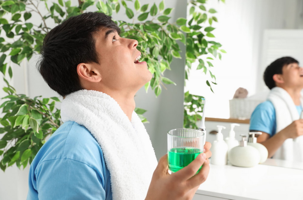 A person in a blue shirt holds a glass of green liquid, surrounded by plants and bathroom products in a well-lit space.