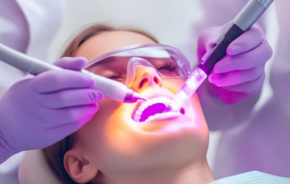 A woman sits in a dental chair while a dentist cleans her teeth with dental tools. 