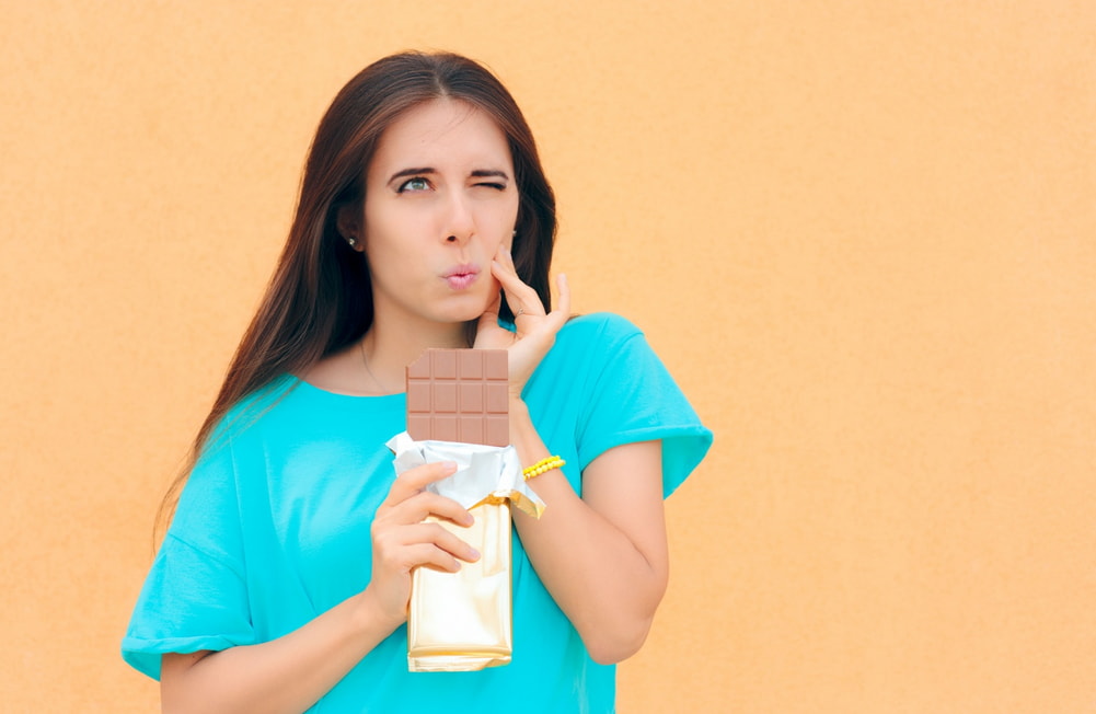 A woman holds a chocolate bar in front of her face, smiling playfully at the camera.