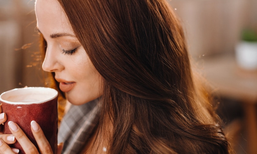 A woman with long hair smiles while holding a steaming cup of coffee in her hands.