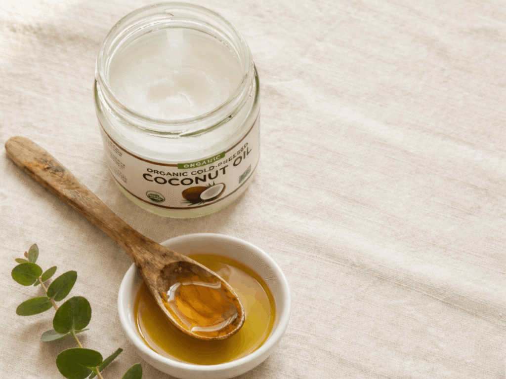 A jar of organic cold-pressed coconut oil beside a wooden spoon and small white bowl of golden oil, with fresh eucalyptus leaves on a linen surface, commonly used for oil pulling for cavities and oral health