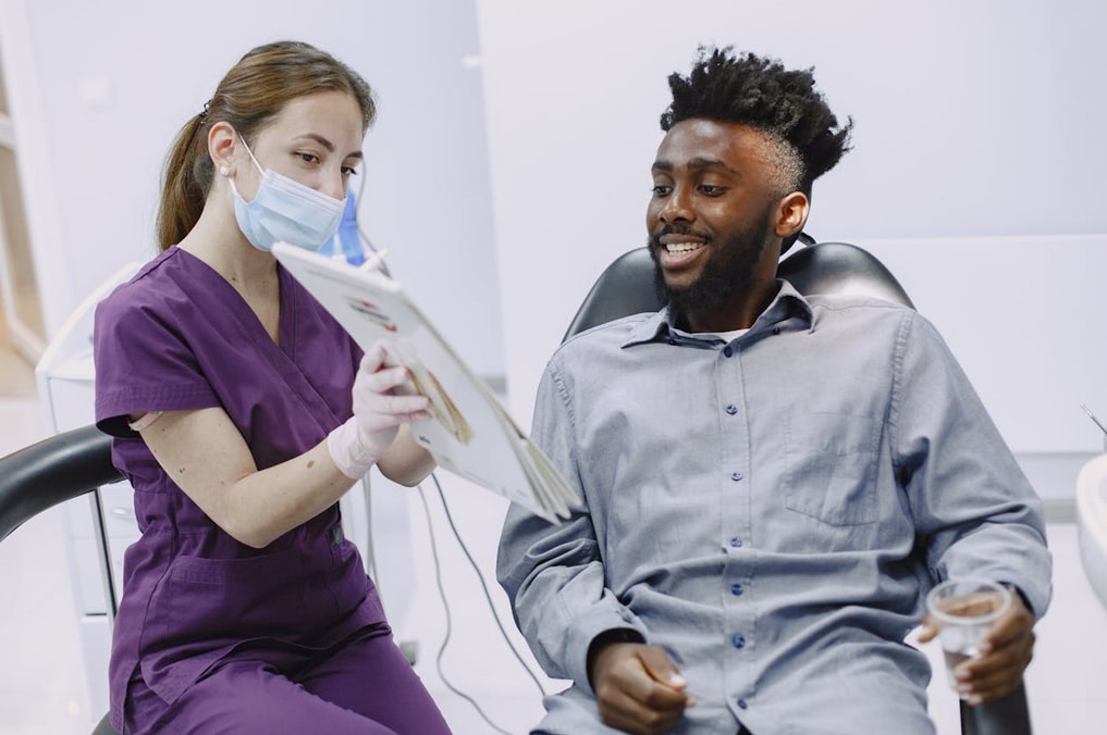 A man and woman seated in dental chairs, discussing dental care with a dentist in a clinical setting.