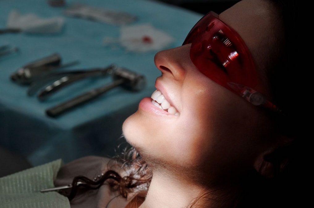 Smiling woman at the dentist wearing red protective glasses, with dental tools on a blue tray in the background.