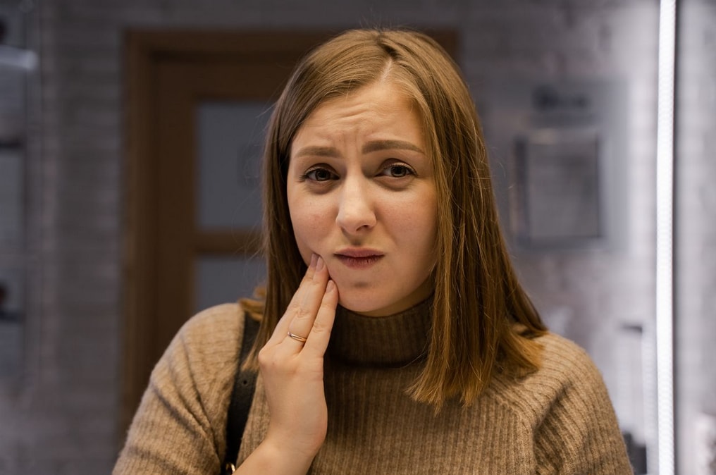 A woman in a brown sweater looks concerned, touching her cheek indoors, possibly experiencing tooth pain or discomfort.