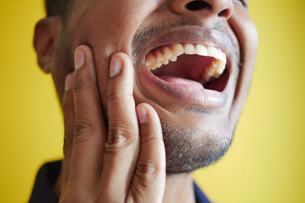 Man with toothache holding cheek, mouth open in pain against a bright yellow background, highlighting dental discomfort.