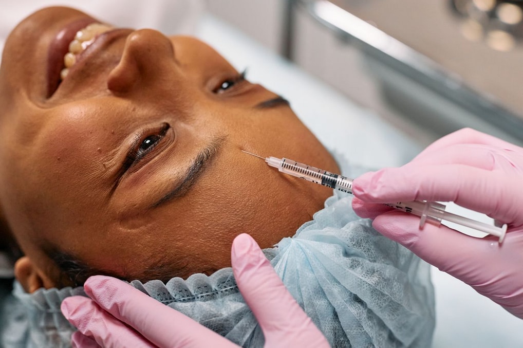 Smiling woman receives a forehead injection from a gloved professional, highlighting a cosmetic procedure in a clinical setting.