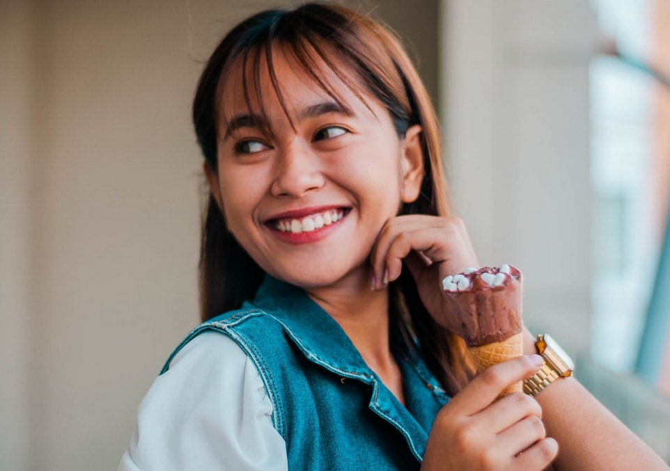 Smiling woman in a denim vest holds a chocolate ice cream cone, looking away joyfully, with a blurred background.
