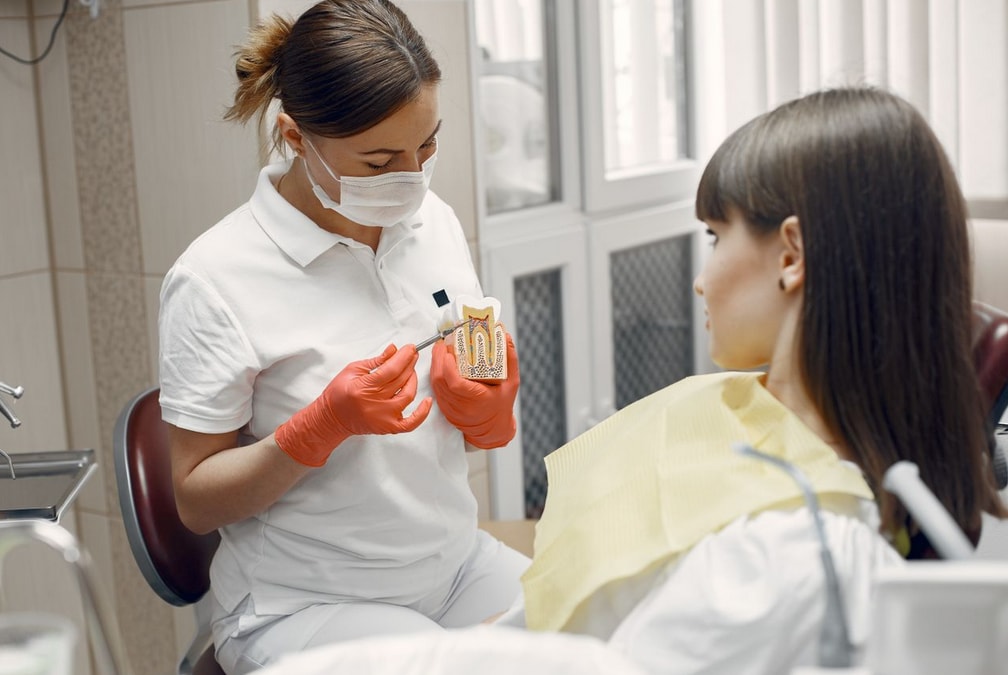 Dentist in mask and gloves shows a tooth model to a seated patient in a clinic, explaining dental care procedures.