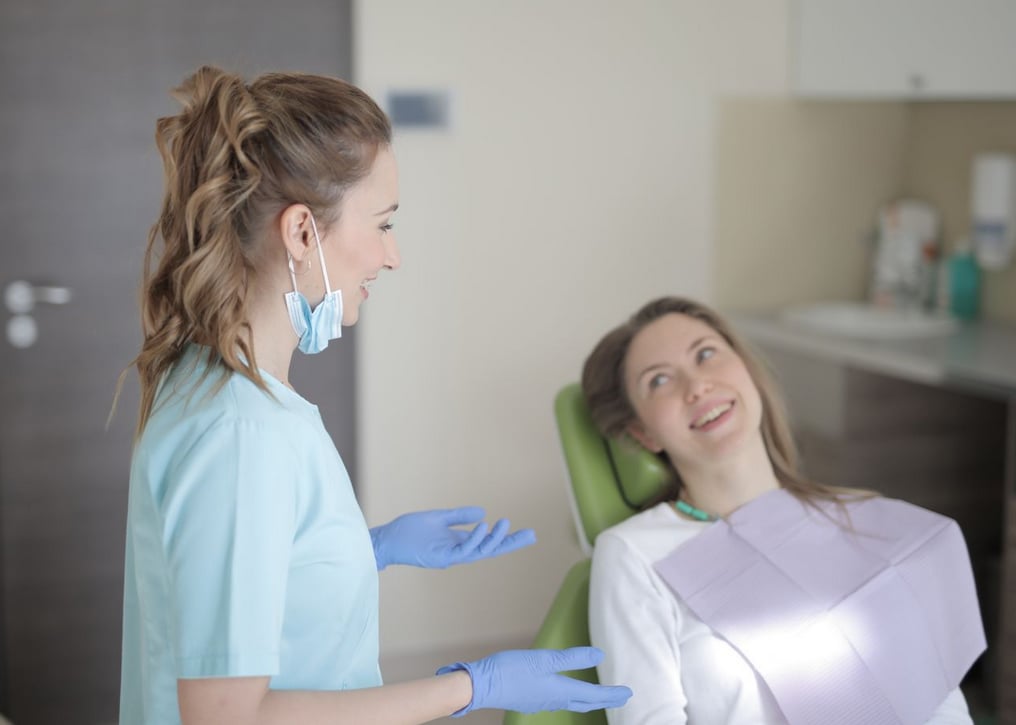 Dentist in blue scrubs and gloves talks to a smiling patient in a dental chair, creating a relaxed and friendly atmosphere.