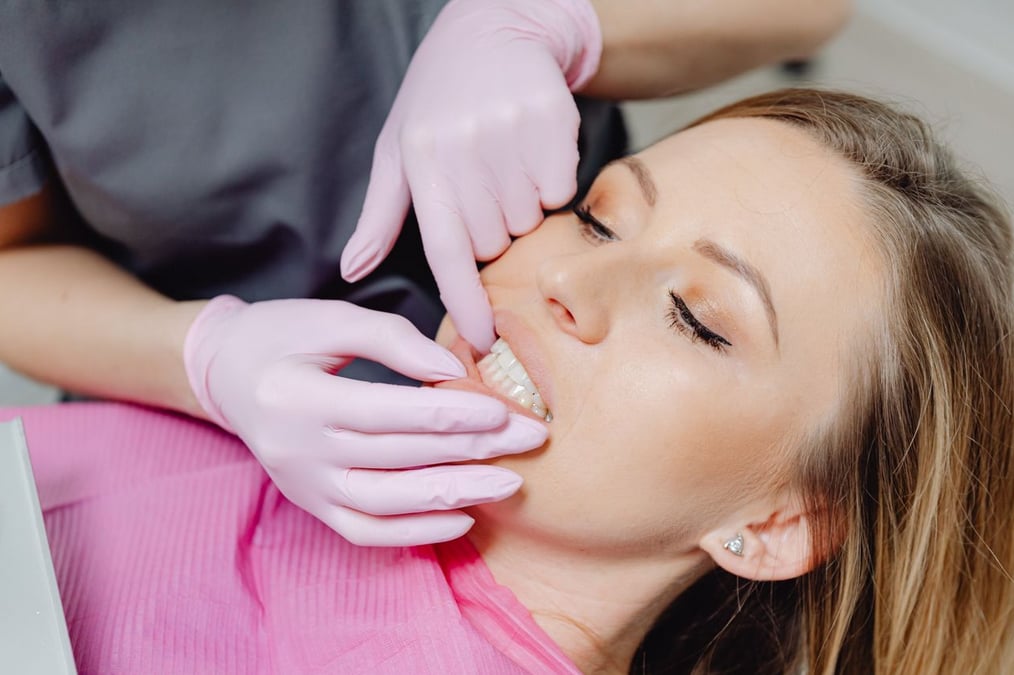 Dentist in pink gloves examines a patient's teeth, focusing on dental health. The patient wears a pink bib, lying back comfortably.