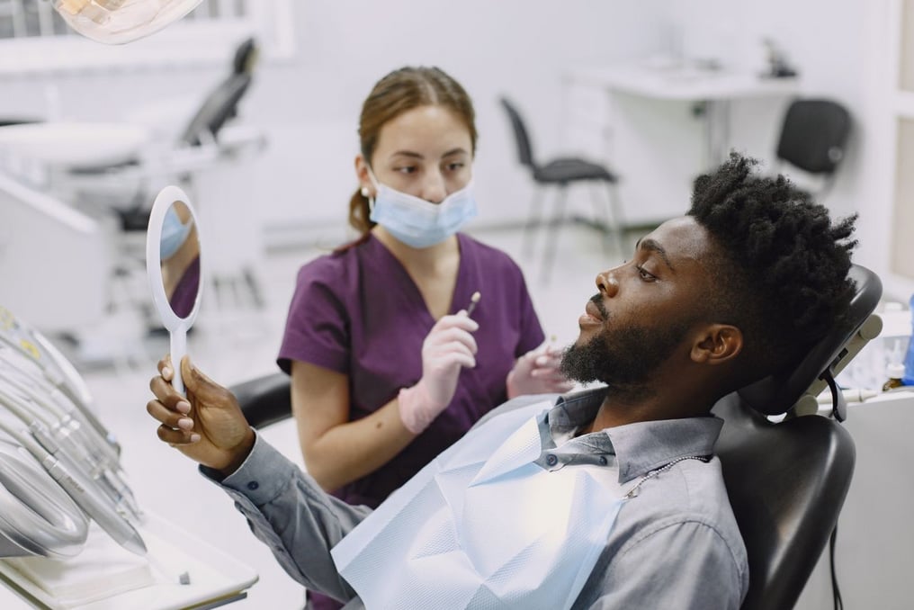 Patient in dental chair holds mirror, examining teeth, while dentist in purple scrubs and mask stands nearby in a bright clinic.