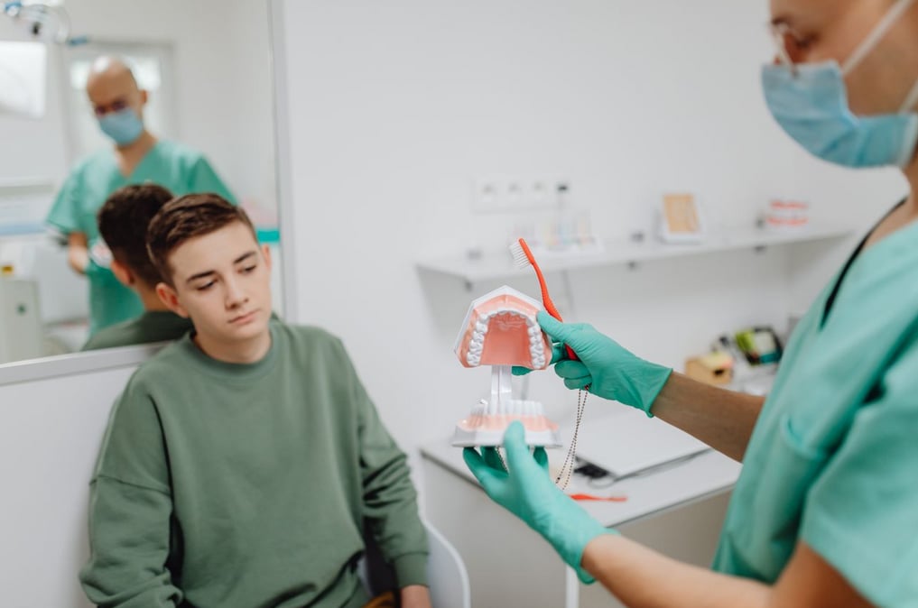Dentist in scrubs shows a dental model to a seated boy, demonstrating brushing technique in a bright clinic room.