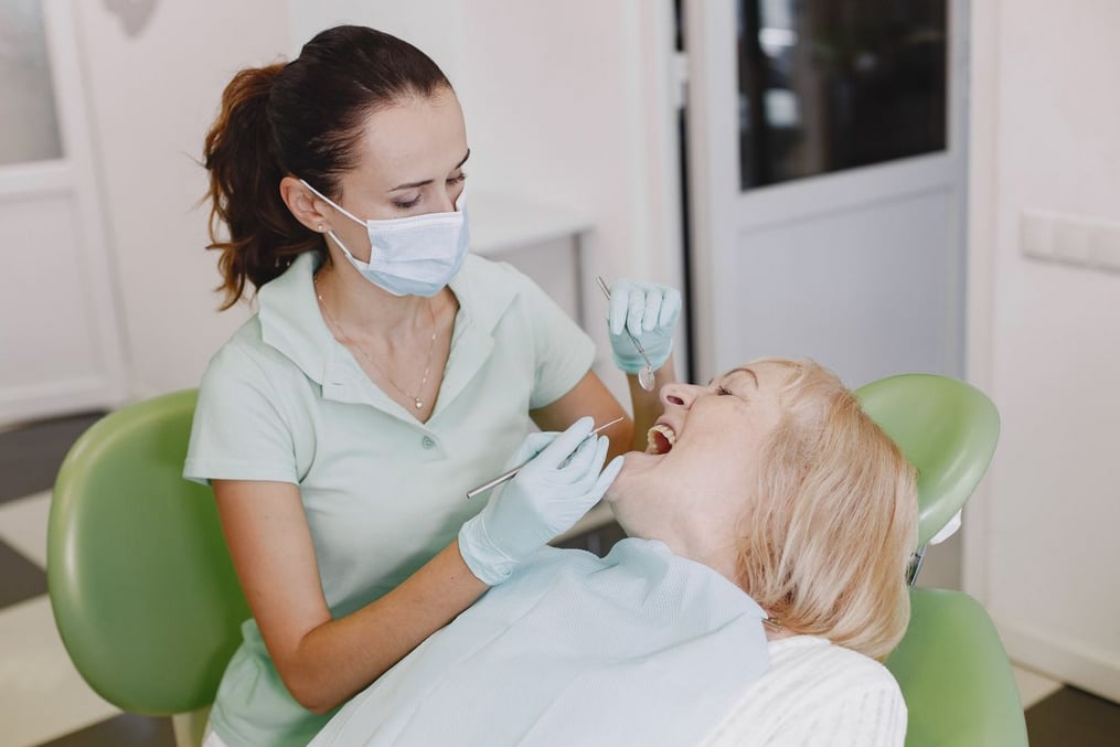 Dentist examines a smiling patient in a dental chair, using tools, in a bright clinic. Both wear protective masks and gloves.