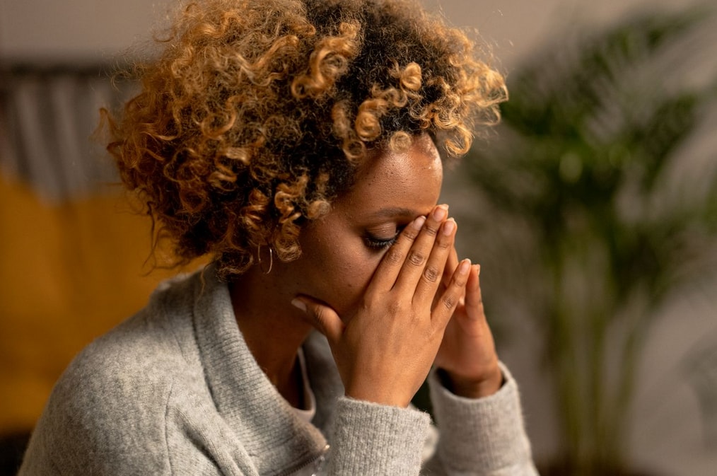 A woman with curly hair sits indoors, hands covering her face, appearing stressed. A plant is blurred in the background.